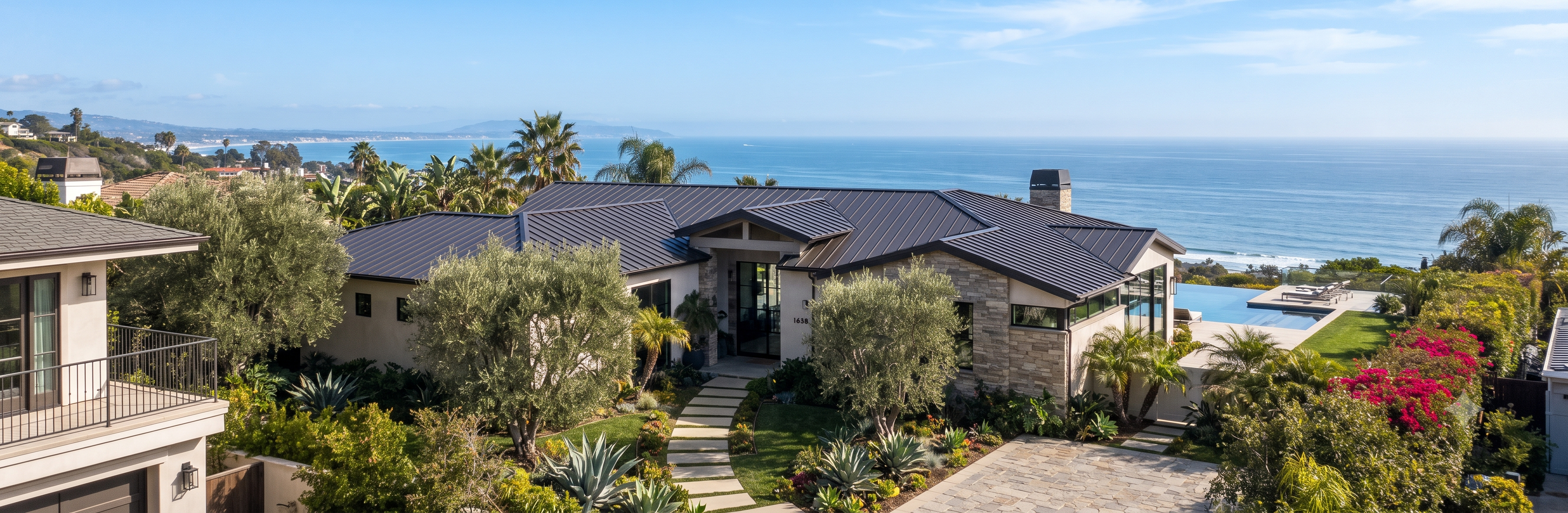 Dark bronze standing seam metal hip roof on a single-story stone-and-stucco estate with lush gardens and ocean views in Pacific Palisades